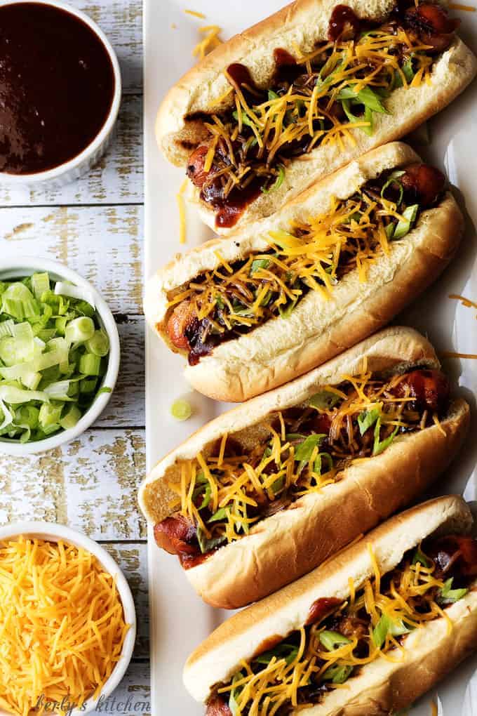 Top down view of four hot dogs on a platter with bowls of cheese, green onions, and bbq sauce.