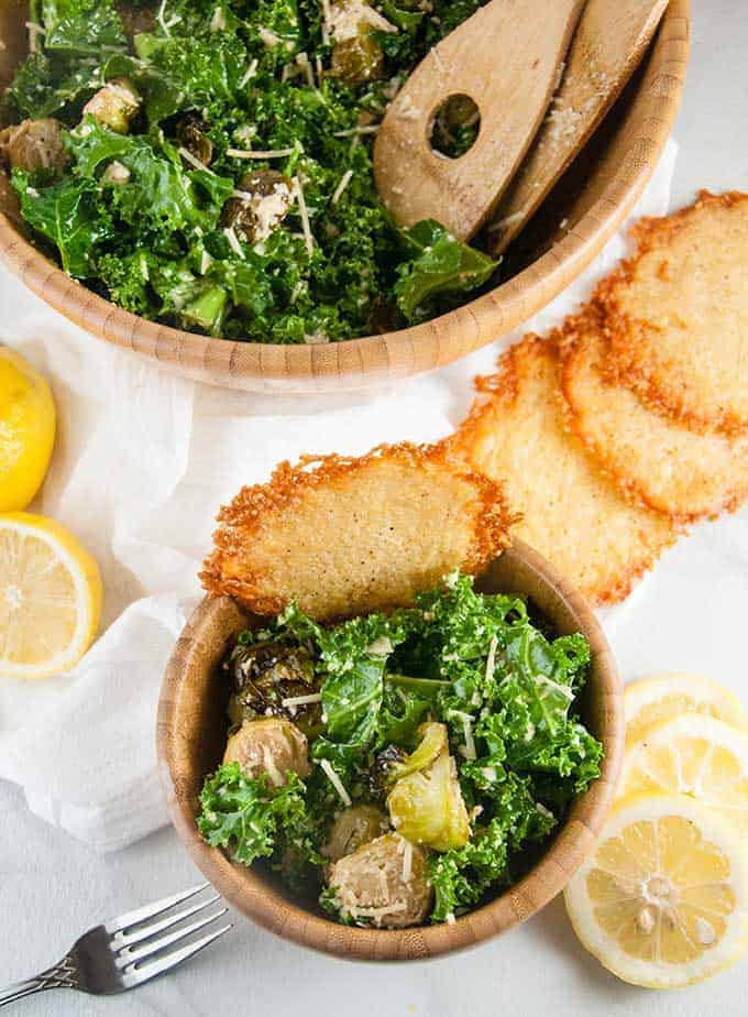 Top down view of a bowl of caesar salad with roasted brussel sprouts next to fresh lemons and parmesan crisps.