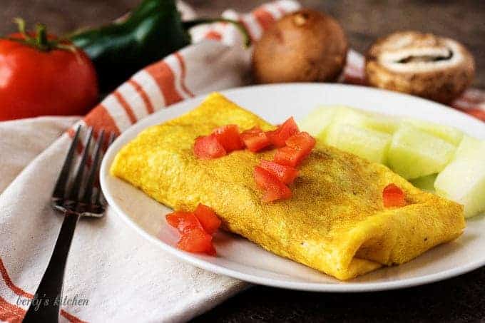 A white plate with the omelet. It's topped with tomatoes and served with melon chunks.