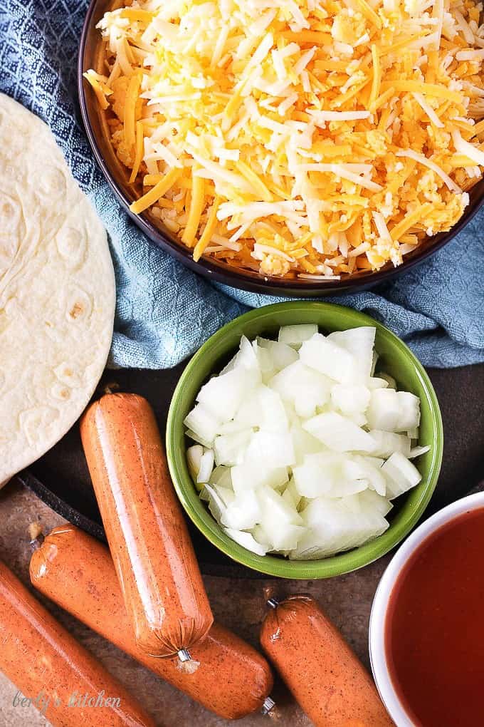 A top-down view of the ingredients, cheddar cheese, onions, sausage in different bowls.