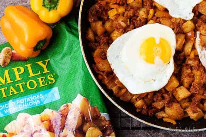 Chorizo hash topped with sunny-side up eggs in a non-stick skillet. Orange sweet peppers and the simply potatoes bag are in the picture.