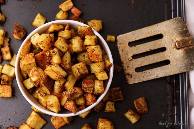 Top-down view of the home fries recipe in bowl, on a sheet pan with a spatula.
