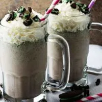 A side-view of two Oreo milkshakes in mugs, topped with whipped cream, and crumbled cookies.