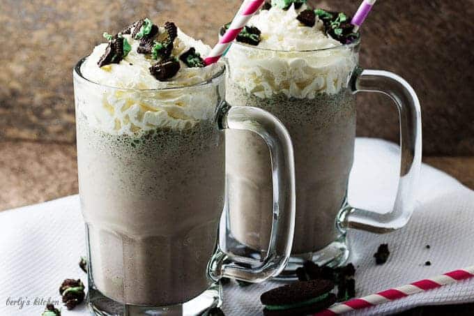 A side-view of two Oreo milkshakes in mugs, topped with whipped cream, and crumbled cookies.
