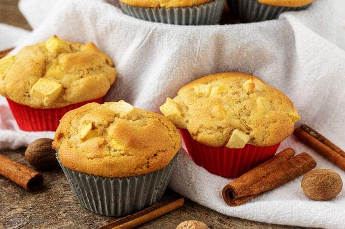 Three finished apple cinnamon muffins in liners in table garnished with cinnamon sticks.