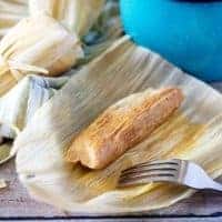 Homemade tamale in an open corn husk next to two tamales and a blue bucket.