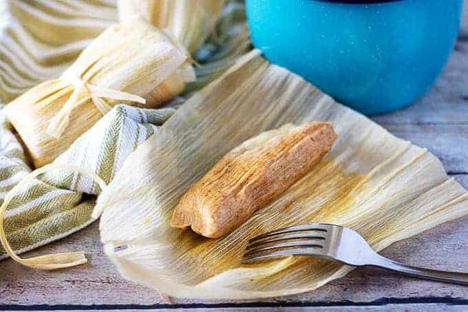 Homemade tamale in an open corn husk next to two tamales and a blue bucket.
