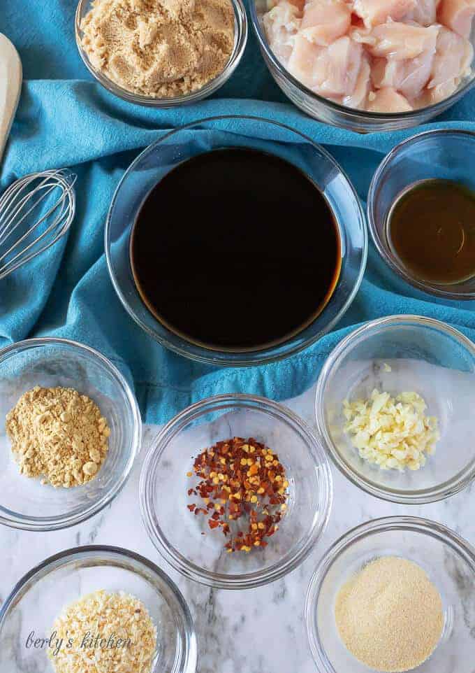 An aerial photo of the ingredients like red pepper flakes, soy sauce, and brown sugar in clear glass bowls.