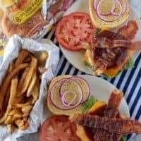 A top-down view of the bacon cheeseburgers being assembled with tomatoes, red onion, and BBQ sauce.