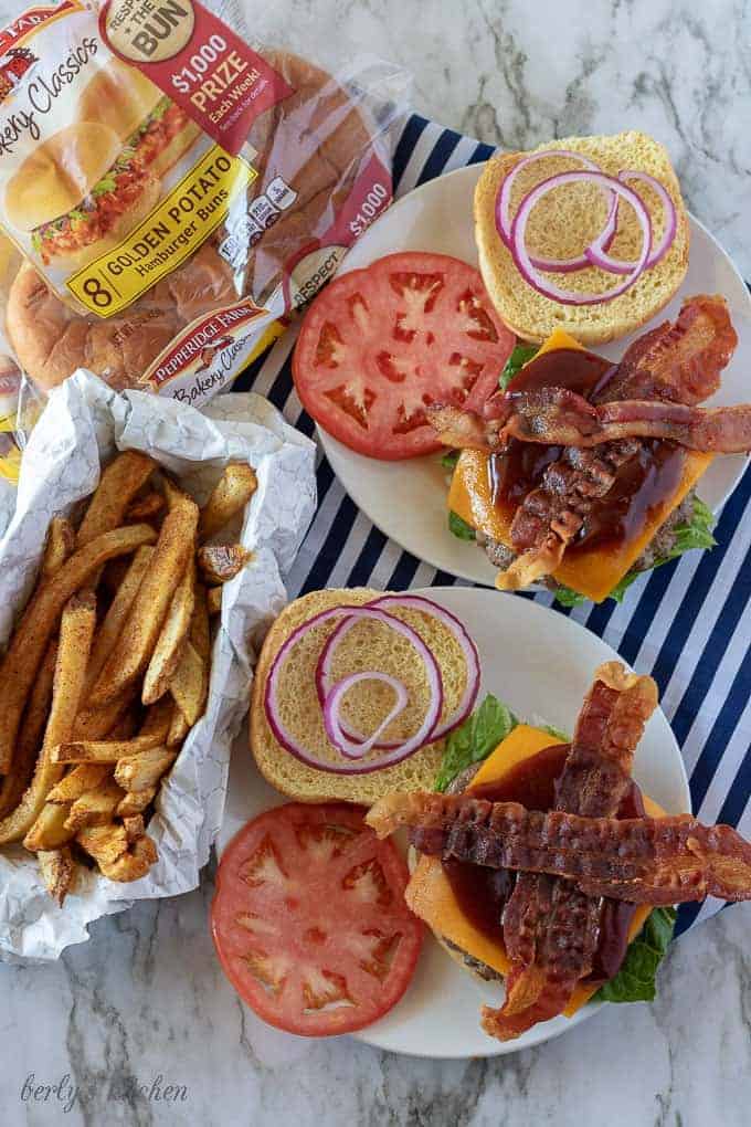 A top-down view of the bacon cheeseburgers being assembled with tomatoes, red onion, and bbq sauce.