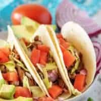 Three shredded beef tacos on a plate next to a tomato and purple onion.