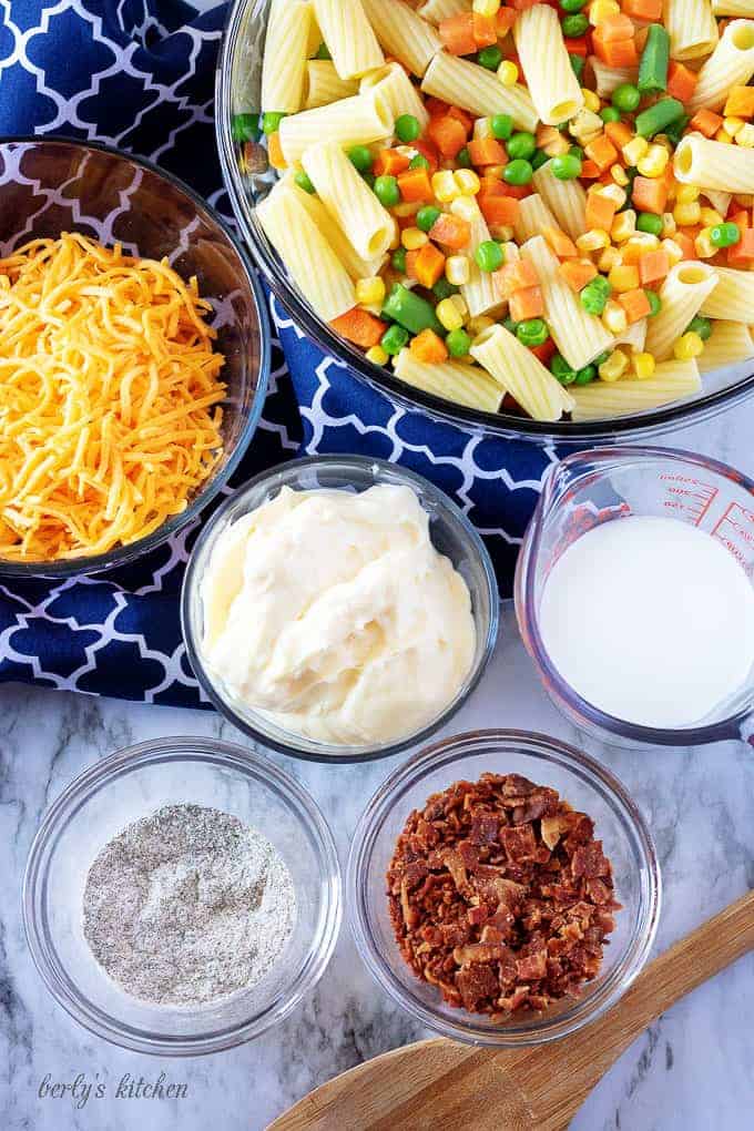 Multiple clear glass mixing bowls showing the ingredients needed for the cold pasta salad, like mayo, bacon, and shredded cheddar cheese.