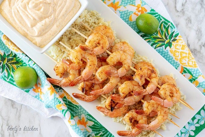 An aerial photo of the finished grilled shrimp on skewers, served over rice, on a rectangular white plate.