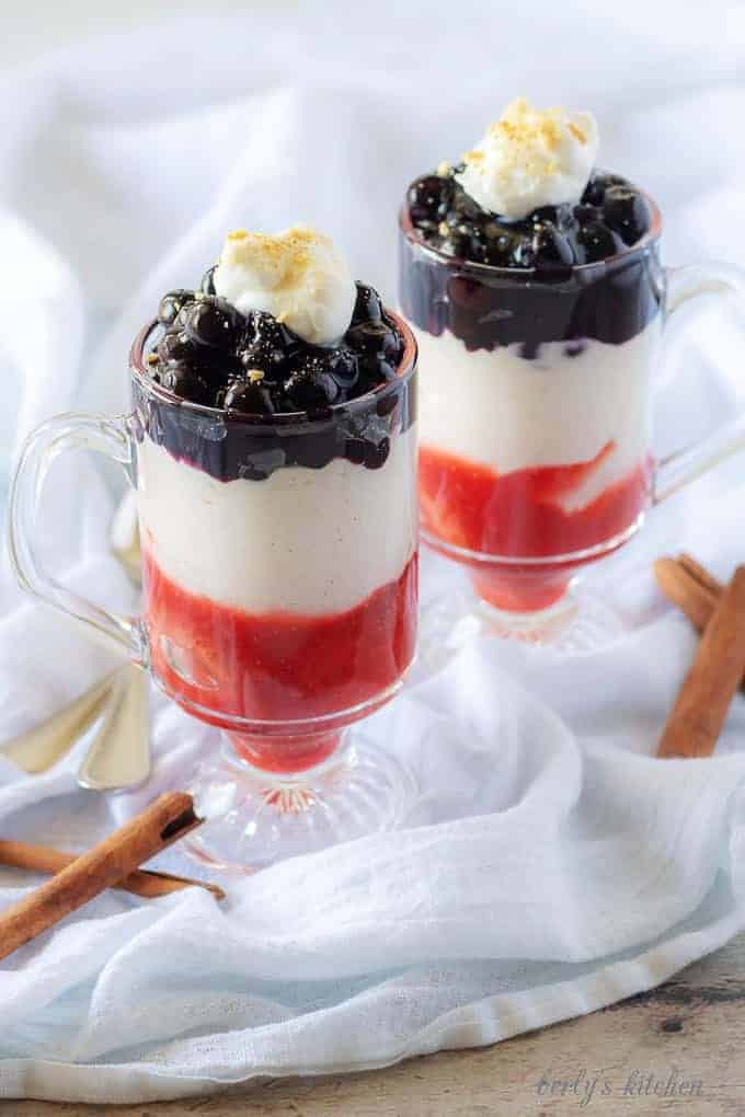 The final picture showing the finished blueberry cheesecake parfaits in clear glass mugs garnished with fresh whipped cream and crumbled graham crackers.