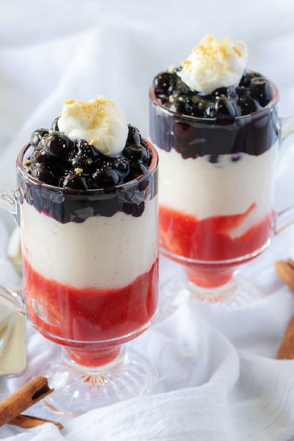 A close-up photo of the finished strawberry blueberry cheesecake parfaits in glass mugs garnished with whipped cream and crumbled graham crackers.