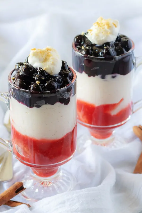A close-up photo of the finished strawberry blueberry cheesecake parfaits in glass mugs garnished with whipped cream and crumbled graham crackers.