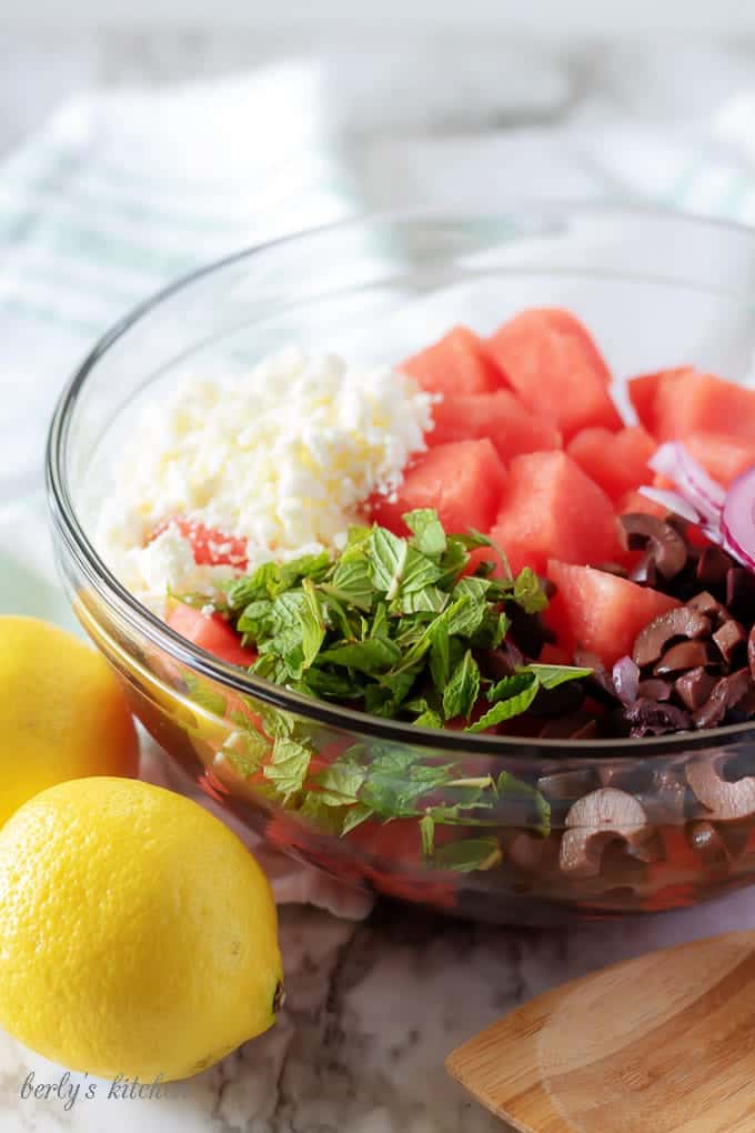 The watermelon salad ingredients, like red onion, fresh mint, feta cheese, and kalamata olives, in a glass mixing bowl.