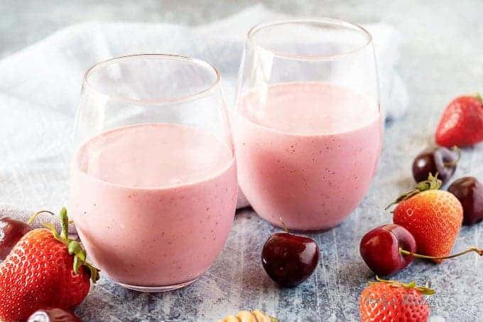 The finished berry smoothies in small glasses on a marble board with cherries and oats sprinkled around them.