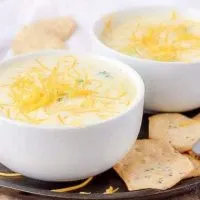 Two bowls of broccoli cheese soup on a flat plate with spoons and crackers.