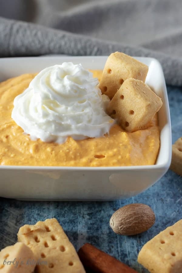 A large picture of the completed pumpkin dip, in a white square bowl, topped with whipped cream, and garnished with a shortbread cookie.