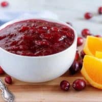 The finished orange cranberry sauce in a white bowl surrounded by fresh cranberries.