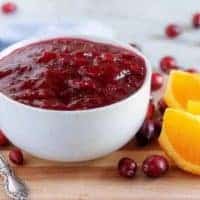 The finished orange cranberry sauce in a white bowl surrounded by fresh cranberries.