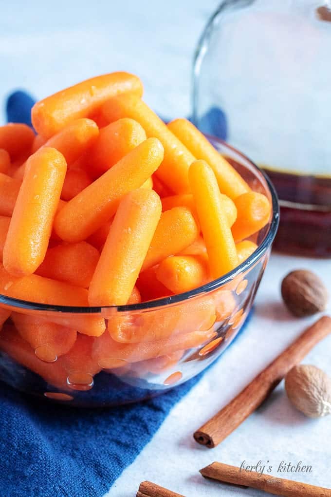 Baby carrots in a glass bowl next to cinnamon sticks, whole nutmeg, and maple syrup.