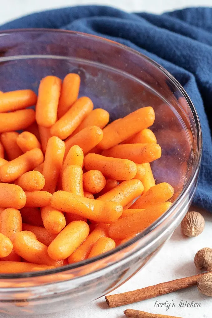 Baby carrots topped with maple syrup, cinnamon, nutmeg, and ginger in a glass bowl.