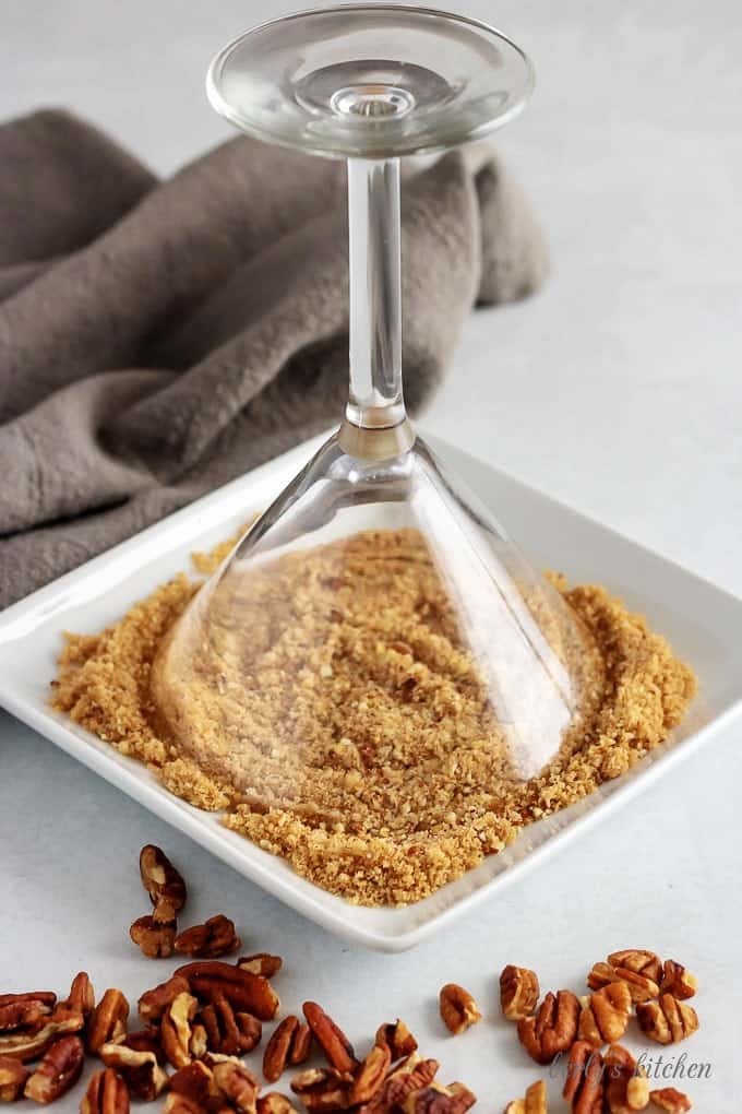 The martini glass has been rimmed with maple syrup and is being dipped into a bowl with crushed pecans and brown sugar.