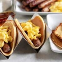 Two breakfast tacos in holders loaded with eggs, sausage, and syrup.