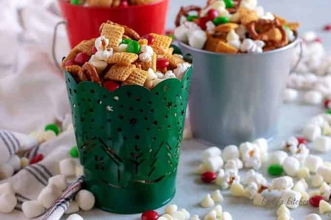 Christmas chex mix in festive tins with popcorn and candy scattered around.