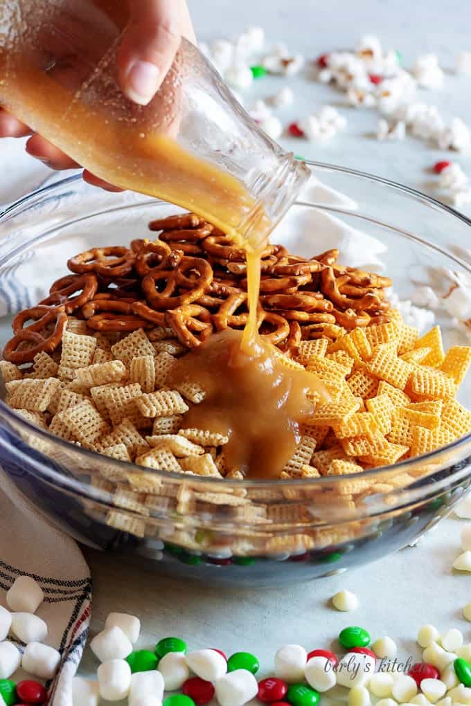 Large bowl of rice chex, corn chex, and mini pretzels being topped with caramel sauce for christmas chex mix.