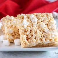 Two perfect rice krispie treats on a square plate next to marshmallows and a red napkin.