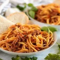 Two pasta bowls filled with beef bolognese and linguine, topped with bread.