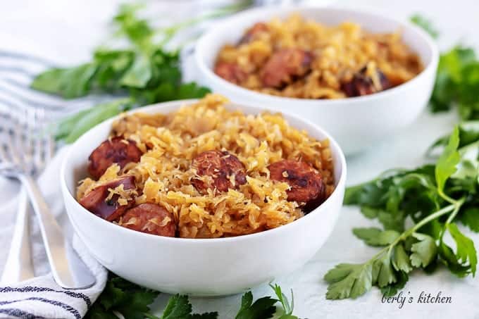 Two bowls of kielbasa and sauerkraut surrounded by fresh parsley.