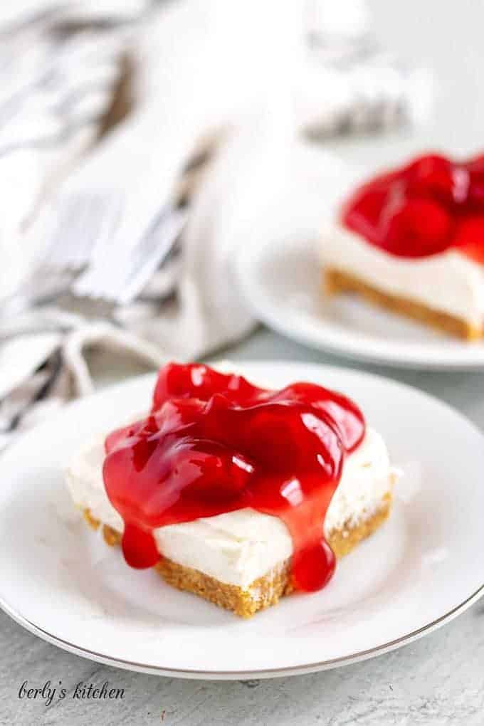 Two cheesecake bars, on white plates, covered with cherry topping.