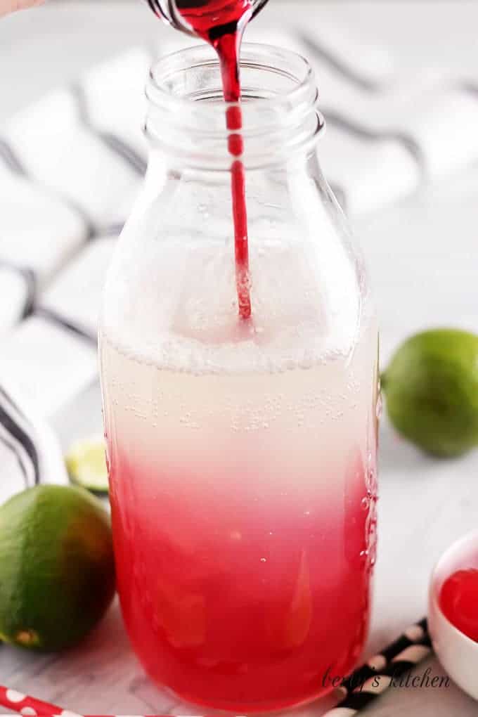 The cherry syrup being poured into the large jar of soda.