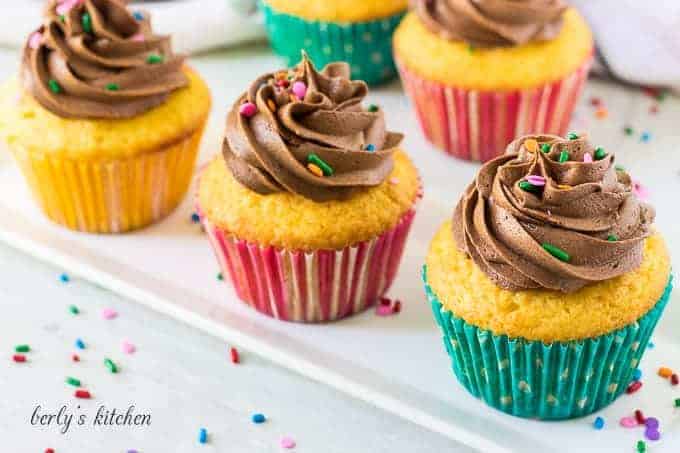 Five cupcakes topped with chocolate buttercream frosting and colorful sprinkles.