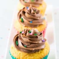 Three yellow cupcakes topped with chocolate buttercream frosting and sprinkles.
