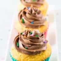 Three yellow cupcakes topped with chocolate buttercream frosting and sprinkles.