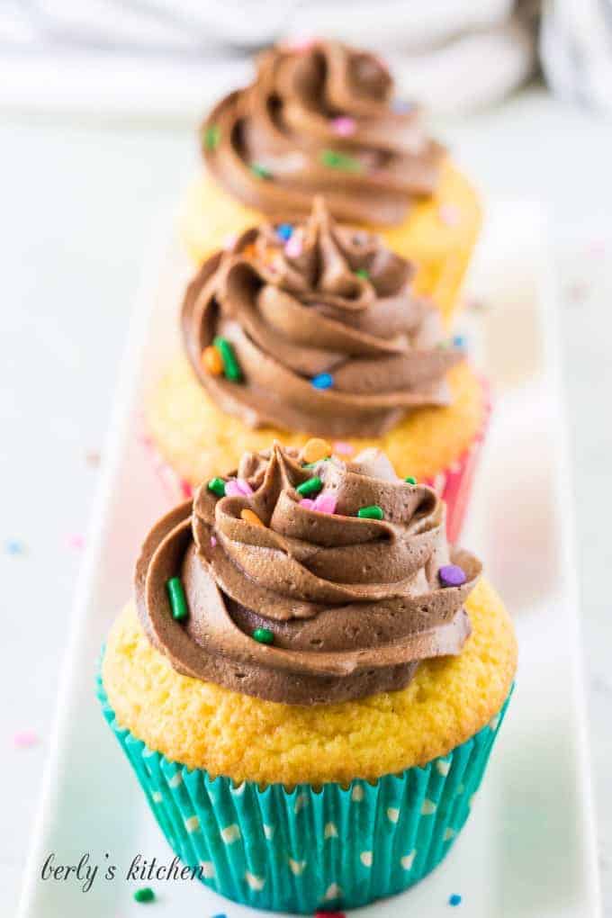Three yellow cupcakes topped with chocolate buttercream frosting and sprinkles.