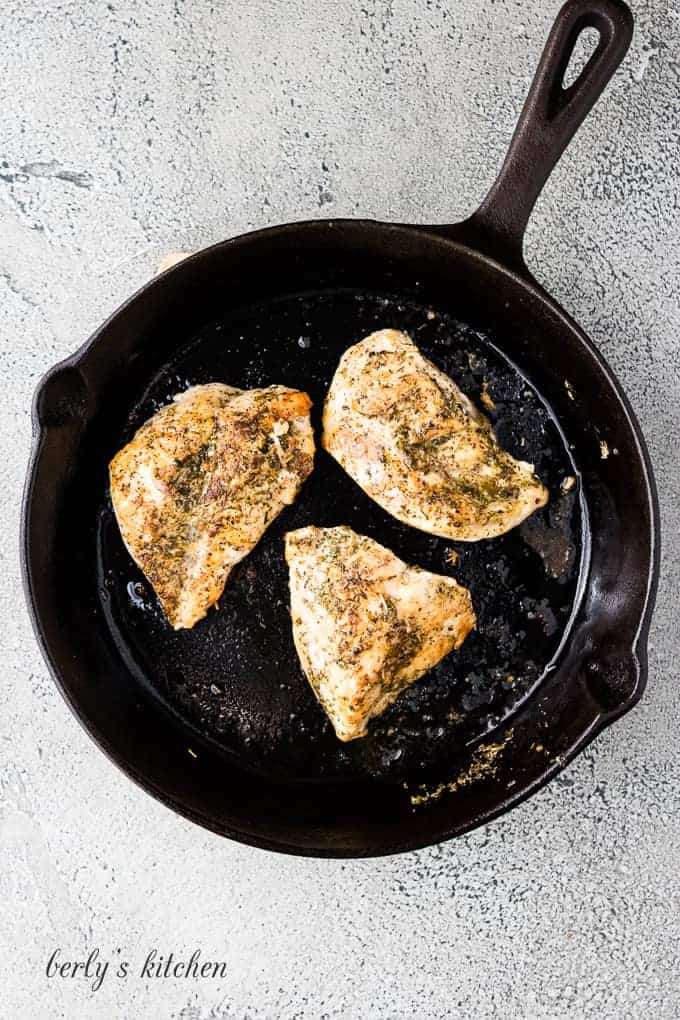 Three seasoned chicken breasts cooking in a cast iron skillet.