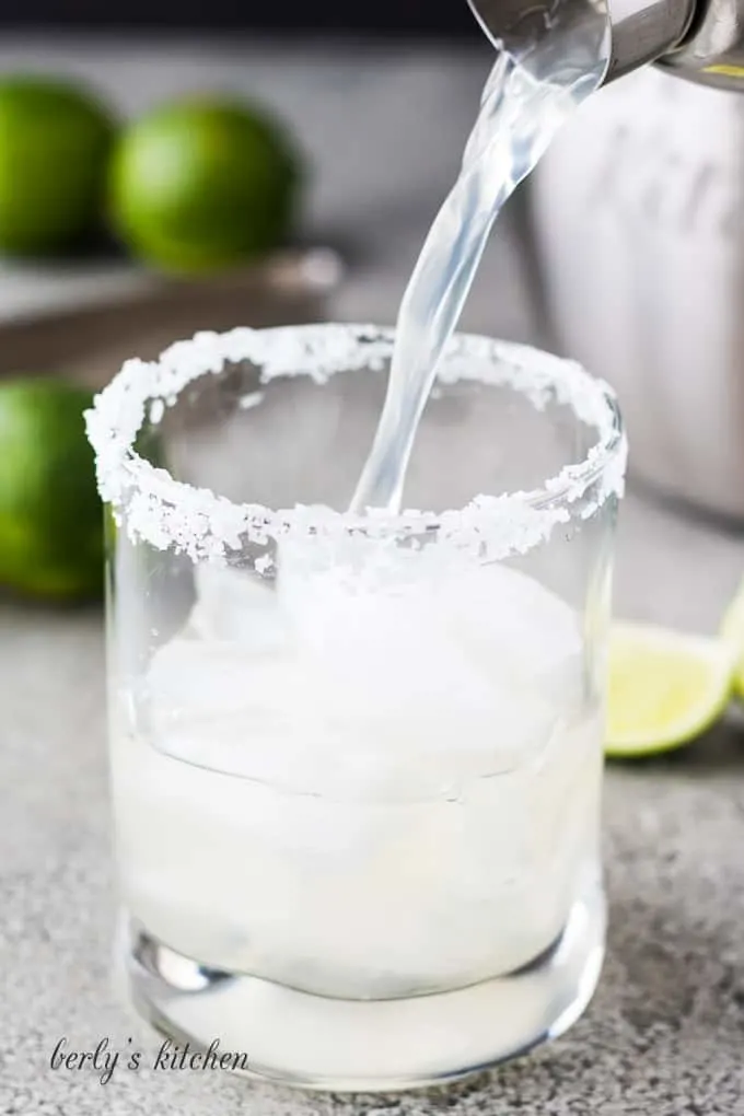 The margarita being poured into the cocktail glass with ice.