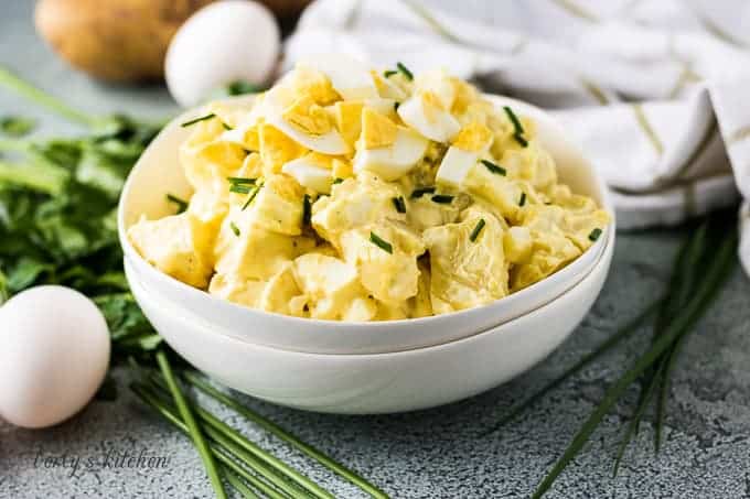 White bowl filled with potato salad next to eggs and chives.