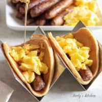 Two pancake breakfast tacos filled with sausage links and scrambled eggs being topped with syrup.