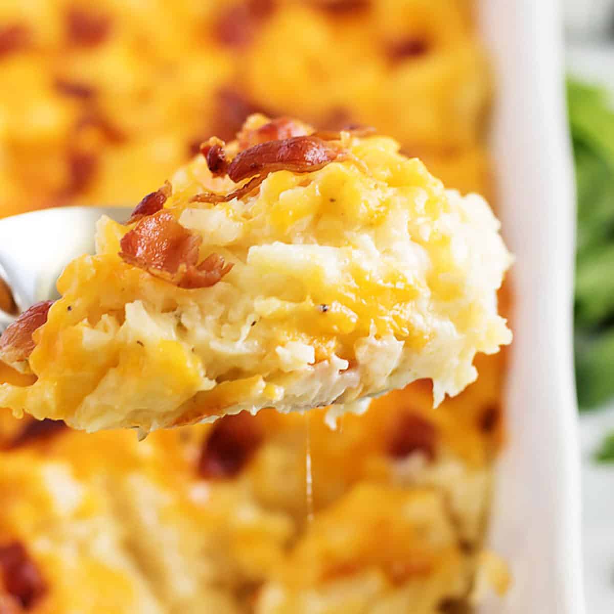 Scoop of hash brown casserole with bacon being held over a baking dish.