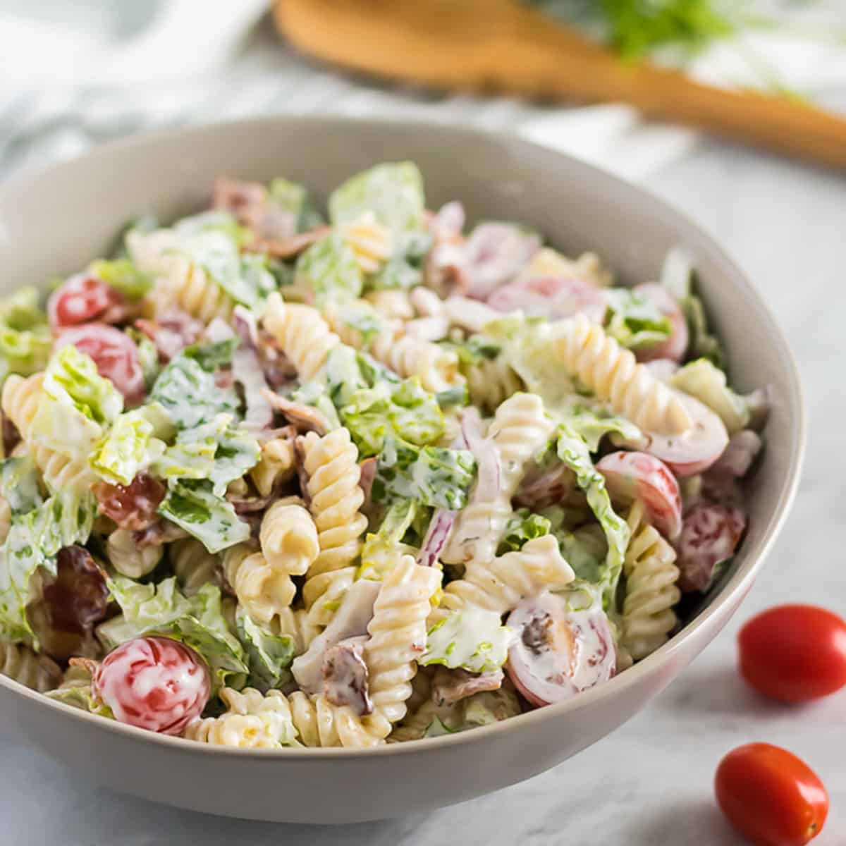 Bowl of BLT pasta salad next to a wooden spoon and tomatoes.
