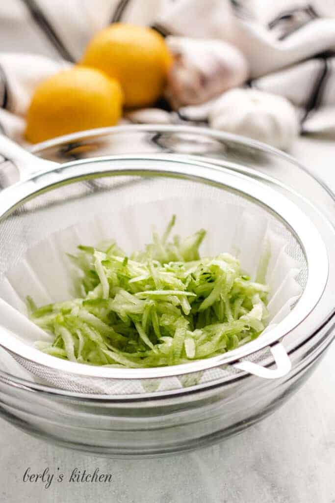 Shredded cucumber in a sieve over a bowl.