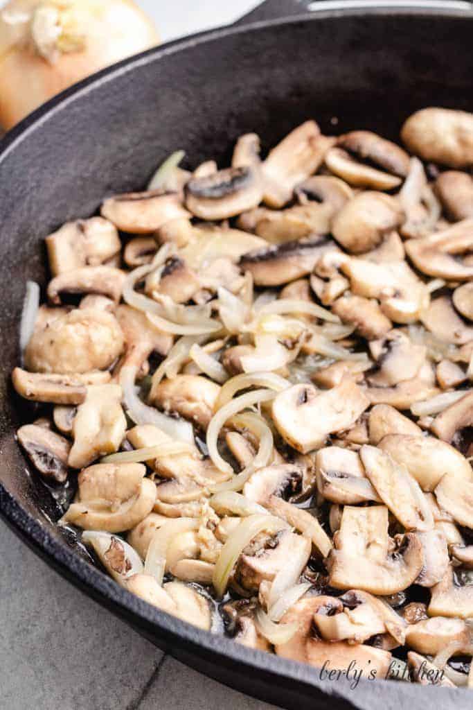 Sliced mushrooms and other ingredients cooking in a skillet.