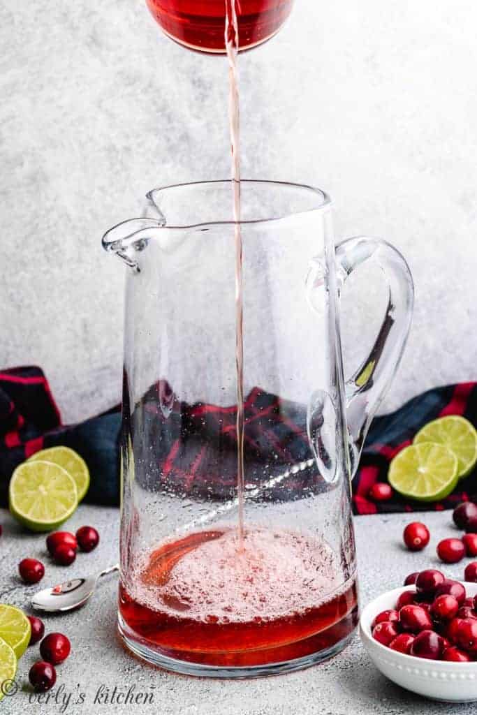 Cranberry juice being added to a large glass pitcher.